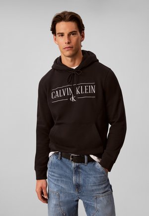 Junger Mann trägt einen schwarzen Calvin Klein Hoodie mit Logo, hellblaue Jeans und schwarzen Gürtel, steht vor einem schlichten grauen Hintergrund.