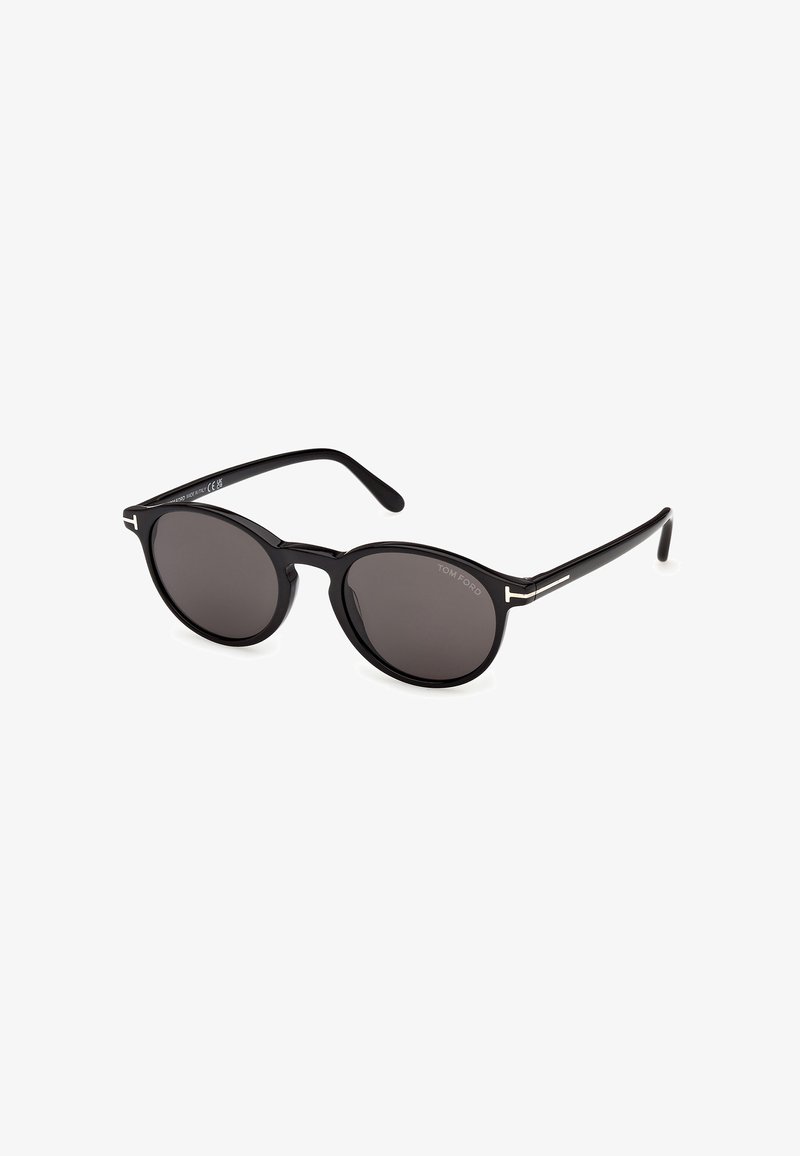 Lunettes de soleil à verres ronds noirs avec montures fines et détails argentés sur les branches, affichant la marque "Tom Ford" sur un verre.