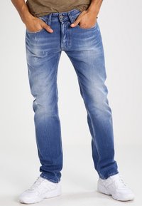 Jeans de mezclilla azul con un acabado desvaído, ligero desgaste, diseño de cinco bolsillos y corte recto. Llevados con zapatillas blancas.