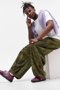 Young man with dreadlocks sits on white block wearing white t-shirt, lavender sweater draped over shoulders, camo pants, and purple sneakers.