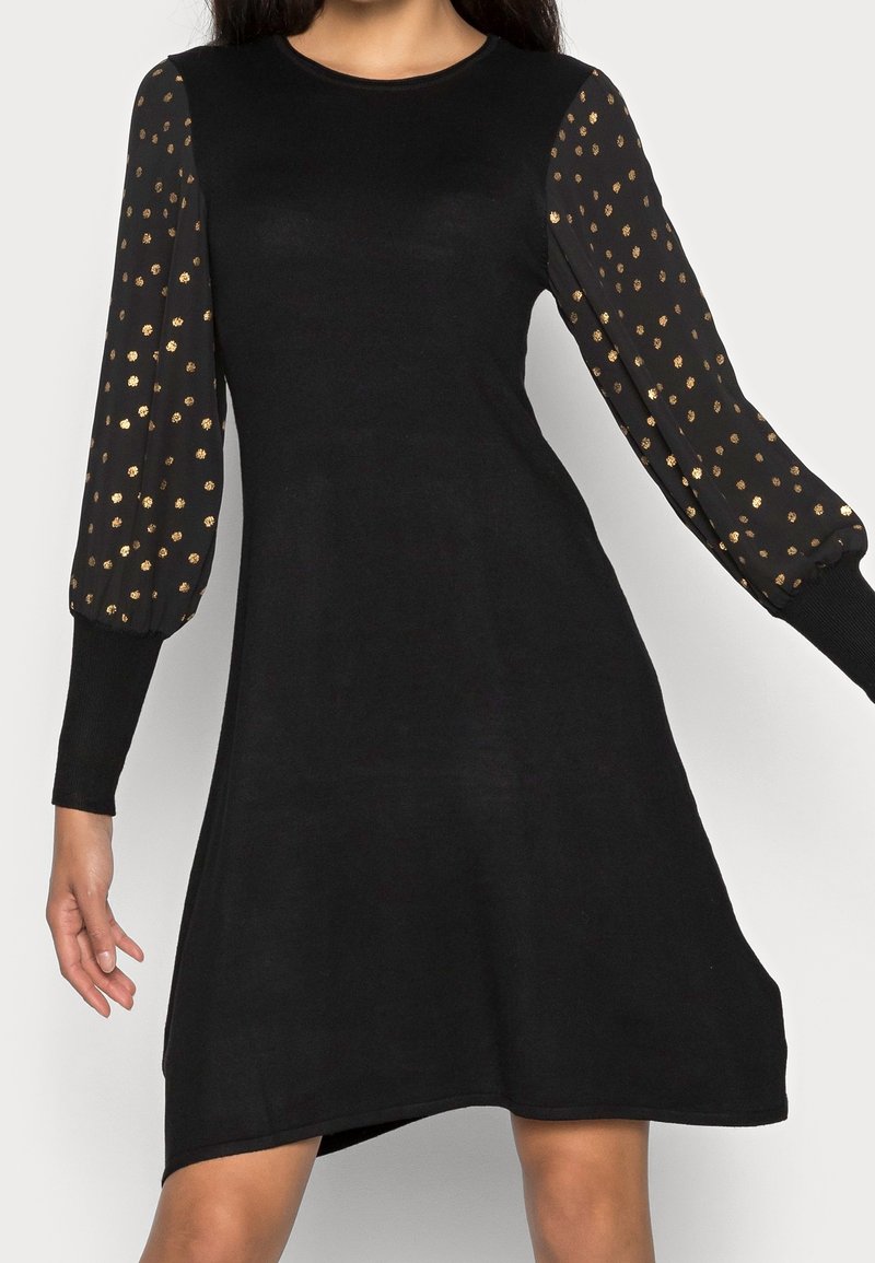 Robe noire en ligne A avec des manches bouffantes noires transparentes ornées de petits pois dorés et des poignets côtelés ajustés.