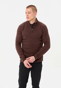 Mladenič v rjavi dolgoročni majici s četrtinskim zadrgo in črnih hlačah, stoji z rokami skupaj, gleda v stran proti enostavnemu ozadju.