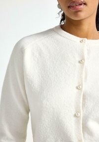 Hvid strikket cardigan med rund halsudskæring, struktureret stof og perleknapper, med raglanærmer og et blødt, hyggeligt udseende.