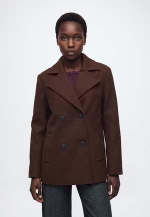 Femme portant un manteau marron foncé croisé avec de larges revers et des boutons noirs, debout devant un fond blanc uni.