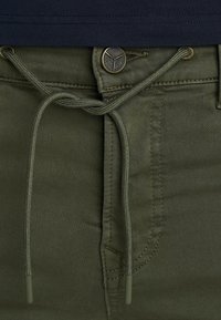 Olijfgroene broek met een elastische tailleband, metalen knoopsluiting en zichtbare stikseldetails. Gladde textuur en aansluitend ontwerp.