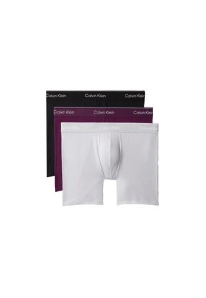 Młody mężczyzna stoi boso na jednolitym tle, nosząc ciemnofioletowe bokserki Calvin Klein.