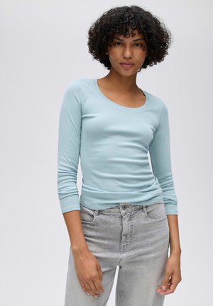 Femme aux cheveux bouclés portant une chemise à manches longues bleu clair et un jean gris clair, debout devant un fond blanc uni.