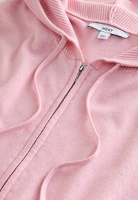 Jasnoróżowa bluza zapinana na zamek, wykonana z miękkiego materiału, z prążkowanymi mankietami i kołnierzem, z zamkiem błyskawicznym i sznurkami. Oznaczona napisem "NEXT". Drobno teksturowana powierzchnia.