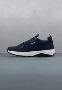 Basket bleu marine avec lacets noirs, semelle blanche, boucle rouge au talon et logo "HUGO" sur le côté, posée sur une surface grise avec un fond gris.
