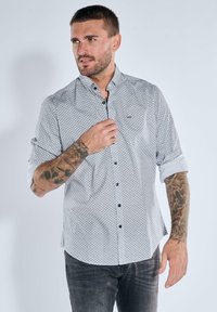 Man with tattoos wearing a white patterned button-up shirt with rolled sleeves and dark jeans, looking to the side.
