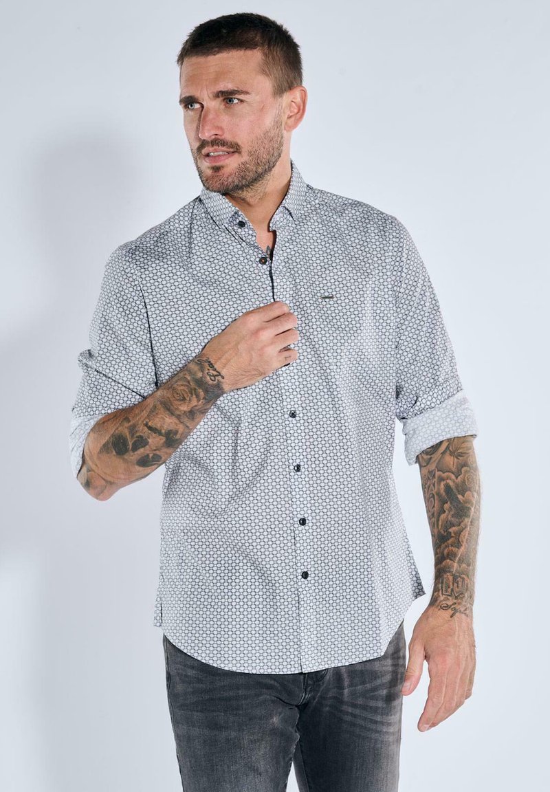 Man with tattoos wearing a white patterned button-up shirt with rolled sleeves and dark jeans, looking to the side.