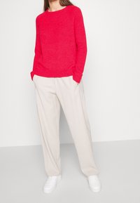 Roter gestrickter Pullover mit rundem Halsausschnitt, gerippten Bündchen und Saum. Kombiniert mit beigen weiten Hosen und weißen Sneakers.