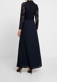 Laulu pituinen laivaston sininen mekko, jossa on pitsinen yläosa, pitkät hihat, korkea kaula-aukko ja sileä hame. Takaa näkyy saumatonta muotoilua ja pitsitietoa.