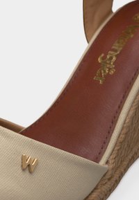 Πλατφόρμα μπεζ σανδάλι με πλεκτή σόλα, καφέ δερμάτινη εσωτερική σόλα με χρυσό λογότυπο και μικρό χρυσό διακοσμητικό "W" στο λουράκι του δακτύλου.