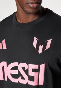 Camiseta de algodón negra con cuello redondo, que presenta un logotipo rosa y detalles de texto. Textura suave con elementos de diseño en contraste.