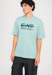Šviesiai melsvas trumpomis rankovėmis t-shirt su apvaliu kaklu, su tamsiai žaliu grafiniu logotipu "EA7" ir "Emporio Armani" priekyje.