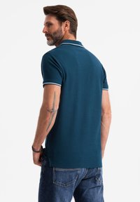 Tyrkysová polokošile s límečkem a bílými akcenty, vyrobená z strukturované látky. Nese ji muž otočený zády, v kombinaci s modrými džíny.