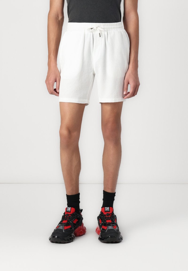 Shorts blancs côtelés avec une taille élastique et un cordon de serrage, associés à des baskets chunky noires et rouges et des chaussettes de cheville.
