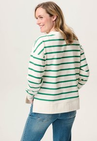 Vrouw die over haar schouder glimlacht, gekleed in een wit en groen gestreepte longsleeve en blauwe spijkerbroek, tegen een effen achtergrond.