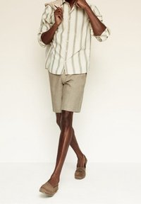 Licht gestreept overhemd met knopen in beige en wit, gecombineerd met beige linnen shorts. Model draagt bruine espadrille-stijl schoenen.