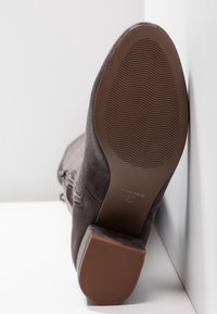 Bottine en daim gris foncé avec un talon bloc, une fermeture éclair latérale visible et une semelle en caoutchouc texturée vue de dessous.