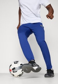 Pantaloni sportivi albaștri, cu un design ajustat și textură din plasă, asortați cu adidași negri, alături de o minge de fotbal cu model.
