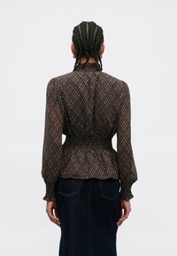 Brun rutete bluse med lange ermer, rynket midje, høy hals og gjennomsiktig stoff. Bakfra vises designdetaljer og passform.
