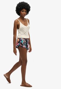 Top de tirantes de punto crema con un patrón de diamantes, combinado con pantalones cortos multicolores con hojas de palma y sandalias de tiras marrones.