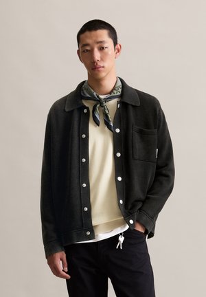 Black, textured button-up jacket with a collar, front pocket, and white buttons layered over a light-colored sweater and dark pants.