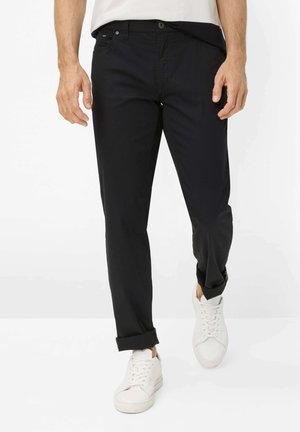 Mand iført sorte opkørte bukser og hvide sneakers, der går fremad mod en ensartet hvid baggrund.