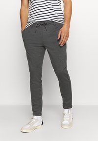 Homme portant un pantalon de jogging gris foncé avec cordon de serrage et poches, associé à des baskets blanches et à une chemise rayée noir et blanc.