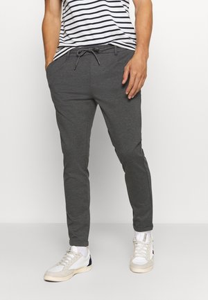 Homme portant un pantalon de jogging gris foncé avec cordon de serrage et poches, associé à des baskets blanches et à une chemise rayée noir et blanc.