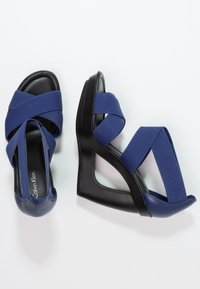 Sandales à talon bleu avec des lanières en tissu croisées, une semelle noire, un bout arrondi et une tige texturée. Nom de la marque visible sur la semelle intérieur.