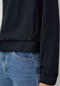 Haut long à manches en satin bleu marine avec un ourlet côtelé, orné d'un accent logo noir rond, associé à un jean en denim bleu clair avec des détails en clous.
