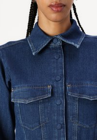 Jeansjack met een donkerblauwe kleur, knopenpatroon aan de voorkant, puntige kraag en twee borstzakken met contrasterende stiksels.