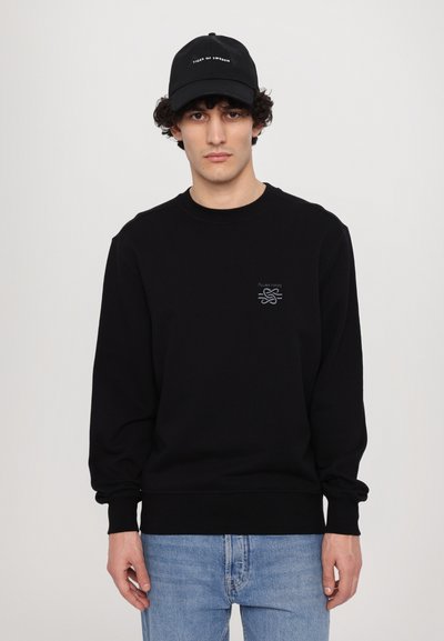 Sweat-shirt noir avec des poignets et un ourlet côtelés, arborant un petit logo brodé sur la poitrine. Associé à un jean bleu clair et une casquette noire.