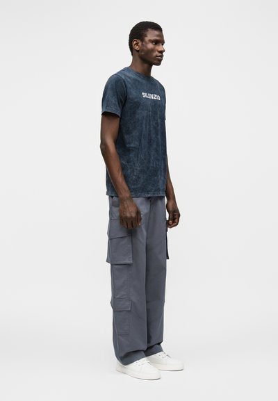 Άνδρας φοράει σκούρο μπλε μπλουζάκι "SILENZIO", γκρι παντελόνι cargo με πολλές τσέπες και λευκά αθλητικά παπούτσια, στέκεται πλαγίως σε απλό φόντο.