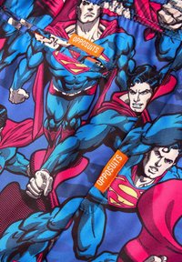 Superherojų tema audinys su ryškiomis mėlynomis, raudonomis ir rožinėmis spalvomis, kuriame pavaizduoti keli spausdinti personažai dinamiškose pozose. Pridėtas „OPPOSUITS“ etiketė.