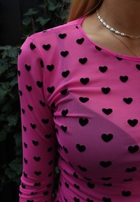 Hosszú ujjú, rózsaszín hálós felső fekete szívmintákkal, kerek nyakkivágással és sima textúrával, finom ezüst ékszerekkel kiegészítve.