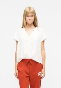 Νεαρή γυναίκα με ίσια ξανθά μαλλιά, που φοράει άνετη λευκή μπλούζα με κοντά μανίκια και κόκκινο παντελόνι φόρμας με κορδόνι και τυπωμένο λογότυπο.