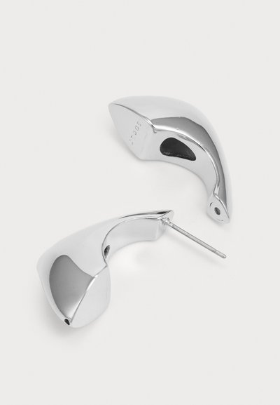 Silverfärgade, skulpturala örhängen med en slät, reflekterande yta och en böjd design. Inkluderar en metallstift för fästning.