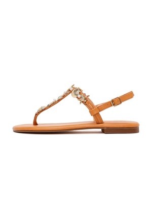 Sandalia plana color canela con correa en el tobillo, diseño de tanga y adornos decorativos dorados de concha marina y perlas en las correas.