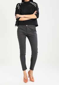 Svart kort topp med vita ränder på axlarna, tillsammans med slim-fit grå byxor. Modellen bär persikofärgade högklackade skor. Minimalistisk design.