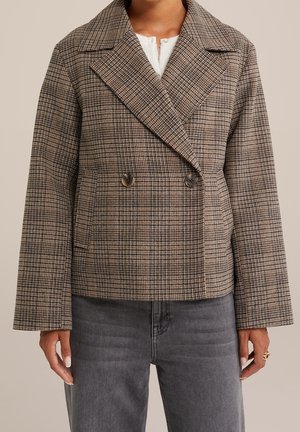 Vrouw draagt een cropped bruin geruit double-breasted blazer met brede revers en donkergrijze spijkerbroek, staat met ontspannen handen.