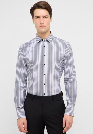 Mand iført en grå og hvid, lodret stribet skjorte med knapper og lange ærmer samt sorte bukser mod en ensfarvet baggrund.