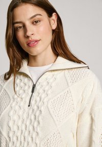 Kreminė megztinė su aukštu apykakle ir pusę užtrauktuku. Papuošta tekstūruotais deimanto ir kabelių raštais, pagaminta iš minkštos vilnos mišinio medžiagos.