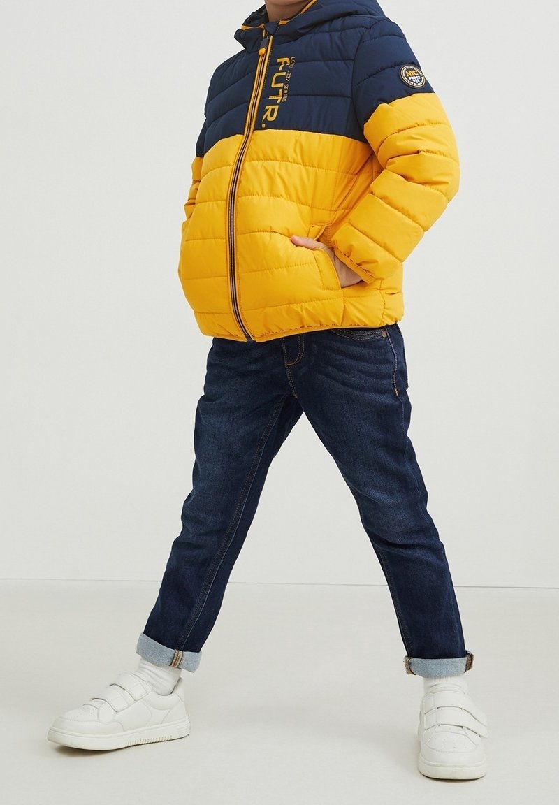 Enfant portant une doudoune jaune et bleu marine, un jean foncé retroussé, des baskets blanches, debout les jambes écartées et une main dans la poche de la veste.