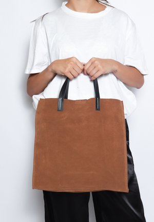 Sac fourre-tout en daim marron avec des poignées en cuir noir, de forme rectangulaire, et une couture soignée. Design simple sans motifs ni accents visibles.