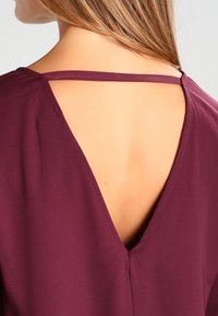 Burgundy topp med V-formad rygg, tunna axelband och en mjuk textur, som framhäver en minimalistisk design utan ytterligare dekorationer.