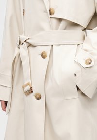 Trench beige avec boutons en écaille, taille ceinturée, détails des boutons aux poignets et petite étiquette drapeau sur la poche, porté par un mannequin.